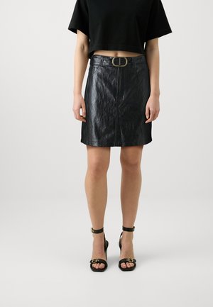 Femme portant un top court noir, une jupe en cuir noire avec ceinture et des sandales à talons noirs, debout devant un fond uni.