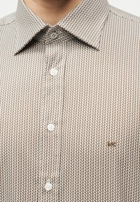 Κοντινό πλάνο σε καφέ και λευκό πουκάμισο με μοτίβο, κουμπωμένο, με μυτερό γιακά και μικρό κεντητό λογότυπο στο στήθος.