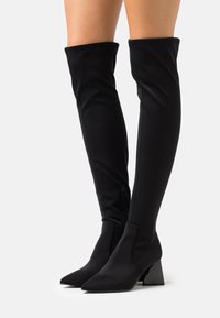 Zwarte laarzen tot boven de dij van rekbaar materiaal, met een spitse neus en een chunky hak. Ritsdetail aan de binnenkant. Gladde textuur.