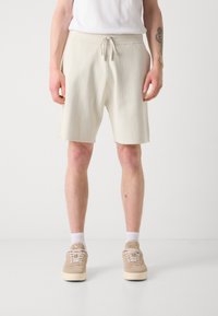Pantalones cortos de color crema con un patrón de rayas verticales texturizadas, cintura con cordón y ajuste relajado, combinados con zapatillas beige y calcetines blancos.