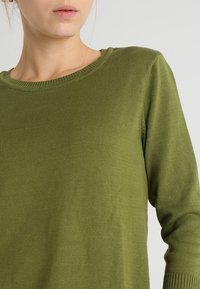 Olivegrüner Strickpullover mit Rundhalsausschnitt und langen Ärmeln mit gerippten Bündchen. Der Stoff ist glatt und hat einen leichten Glanz.