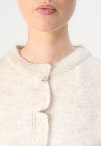 Cardigan lavorato a maglia color beige chiaro con scollatura rotonda, cinque bottoni sul davanti e una texture morbida con sottili variazioni di colore.