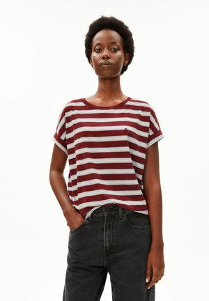 Frau mit kurzen lockigen Haaren, die ein rot-weiß gestreiftes kurzärmeliges Shirt und schwarze Jeans trägt, mit einer Hand in der Tasche.