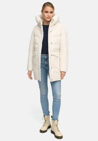 Doudoune blanche avec col montant, fermeture éclair à l'avant et deux grandes poches avant. Assortie à un jean bleu clair et des bottes beiges à lacets.