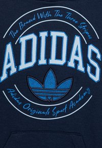 Blå och marinblå hoodie med en broderad Adidas-logotyp, cirkulär textdesign och ett tre-stripes accentemblem nederst.