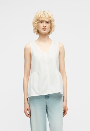 Blond kvinne med krøllete hår iført ermeløs hvit V-hals topp og lys blå høyhulte jeans mot ensfarget lyst bakgrunn.