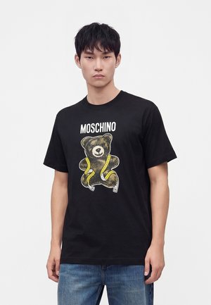 Νέος άντρας που φοράει μαύρο t-shirt Moschino με τυπωμένη στην μπροστινή πλευρά μια αρκούδα τυλιγμένη με κίτρινη μεζούρα.