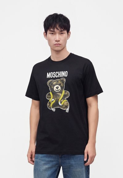 Νέος άντρας που φοράει μαύρο t-shirt Moschino με τυπωμένη στην μπροστινή πλευρά μια αρκούδα τυλιγμένη με κίτρινη μεζούρα.