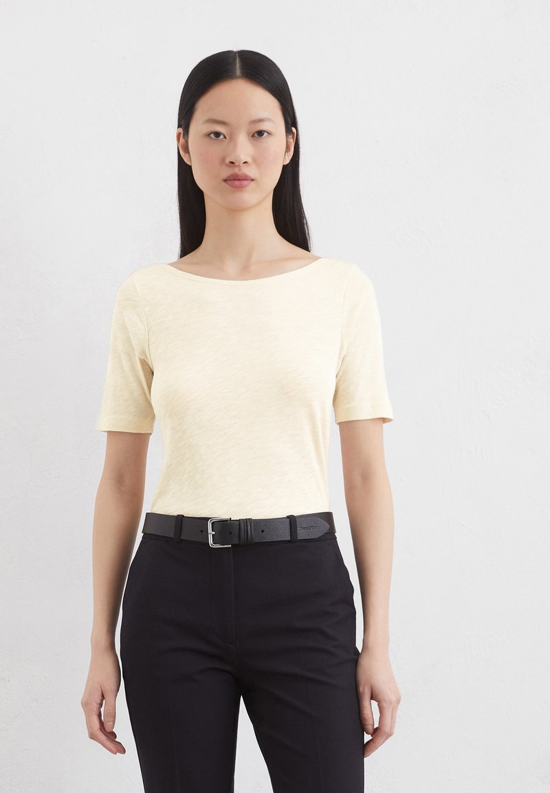 Kurzärmliges, blassgelbes, strukturiertes Oberteil mit weitem U-Boot-Ausschnitt, kombiniert mit dunklen Hosen und einem schwarzen Ledergürtel mit einer silbernen Schnalle.