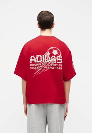Osoba ubrana w czerwoną, za dużą koszulkę Adidas z białą grafiką piłki nożnej i napisem na plecach, w połączeniu z jasnoszarymi spodniami.