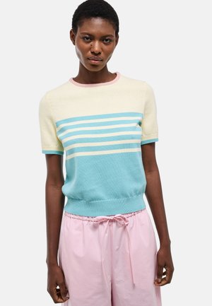 Femme portant un pull à manches courtes à rayures bleues et blanches, associé à un pantalon à cordon de serrage rose clair, sur un fond uni.