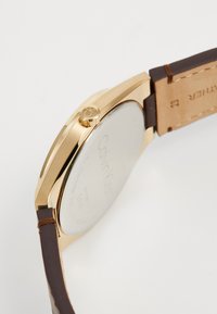 Reloj dorado con caja de acero inoxidable, correa de cuero marrón y una corona prominente que presenta el logo de Calvin Klein.