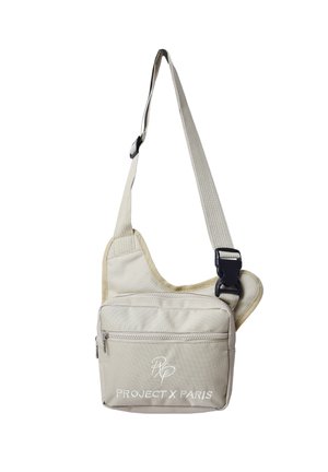 Bolso beige de bandolera con correa ajustable, bolsillo frontal con cremallera y "Project X Paris" escrito en la parte delantera en bordado blanco.