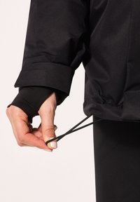 Veste noire en tissu texturé avec des poignets retroussés ; une main tire sur un cordon noir au bas, ajustant son ajustement.