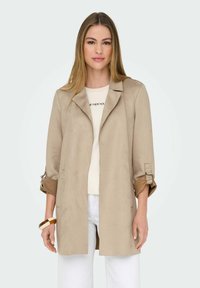 Donna che indossa un blazer lungo beige con maniche arrotolate, una camicia chiara con testo, pantaloni bianchi e un bracciale marrone e crema, in piedi contro uno sfondo bianco.