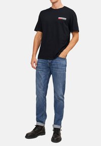 Schwarzes Baumwoll-T-Shirt mit kleinem Logo auf der Brust, kombiniert mit hellblauen Denim-Jeans und schwarzen Schnürstiefeln mit umgeschlagenem Saum.