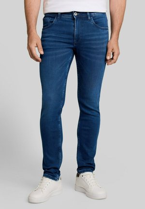 Man die een slim-fit blauwe spijkerbroek en witte sneakers draagt, staand tegen een effen lichtgrijze achtergrond.