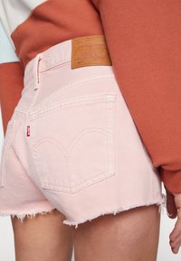 Mujer llevando pantalones cortos de mezclilla deshilachados de color rosa claro con parche de cuero de Levi's y una blusa de manga larga color óxido, mostrada desde atrás.