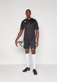 Sort atletisk skjorte og shorts, hvidt logo, parret med sorte fodboldsko og hvide knæhøje sokker, mens han/hun holder en sort og hvid fodbolde.