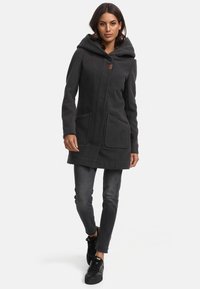 Abrigo con capucha de color gris oscuro, con cierre de cremallera, dos bolsillos frontales y una textura suave. Combinado con jeans oscuros y zapatillas negras.
