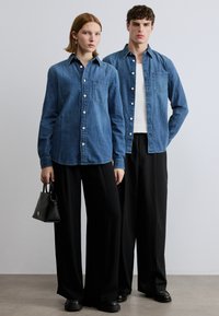 Jeans srajce v srednje modri barvi z gumbi, ena z žepom; kombinirane z ohlapnimi črnimi hlačami in majhno črno torbico.