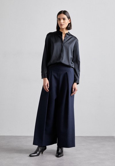 Chemise noire en satin à boutons, associée à un pantalon large bleu marine et des bottes à talons noires. La texture lisse et la coupe ample mettent en avant un design moderne.