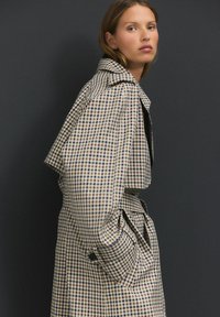 Überprüfter Trenchcoat mit einem beigen und marineblauen Muster, ausgestattet mit breiten Revers, tailliertem Gürtel und seitlichen Taschen. Hergestellt aus strukturiertem Stoff.