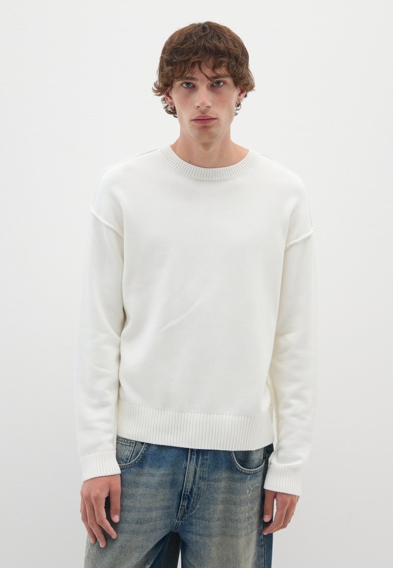 Weißer Strickpullover mit runder Ausschnitt, geripptem Saum und Bündchen. Hat eine lockere Passform und eine glatte Textur, kombiniert mit hellblauen Jeans.