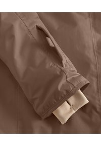 Manche de veste imperméable marron avec un poignet élastique beige doux et une poche angulée fixée par un détail à onglet. Texture lisse, design minimal.