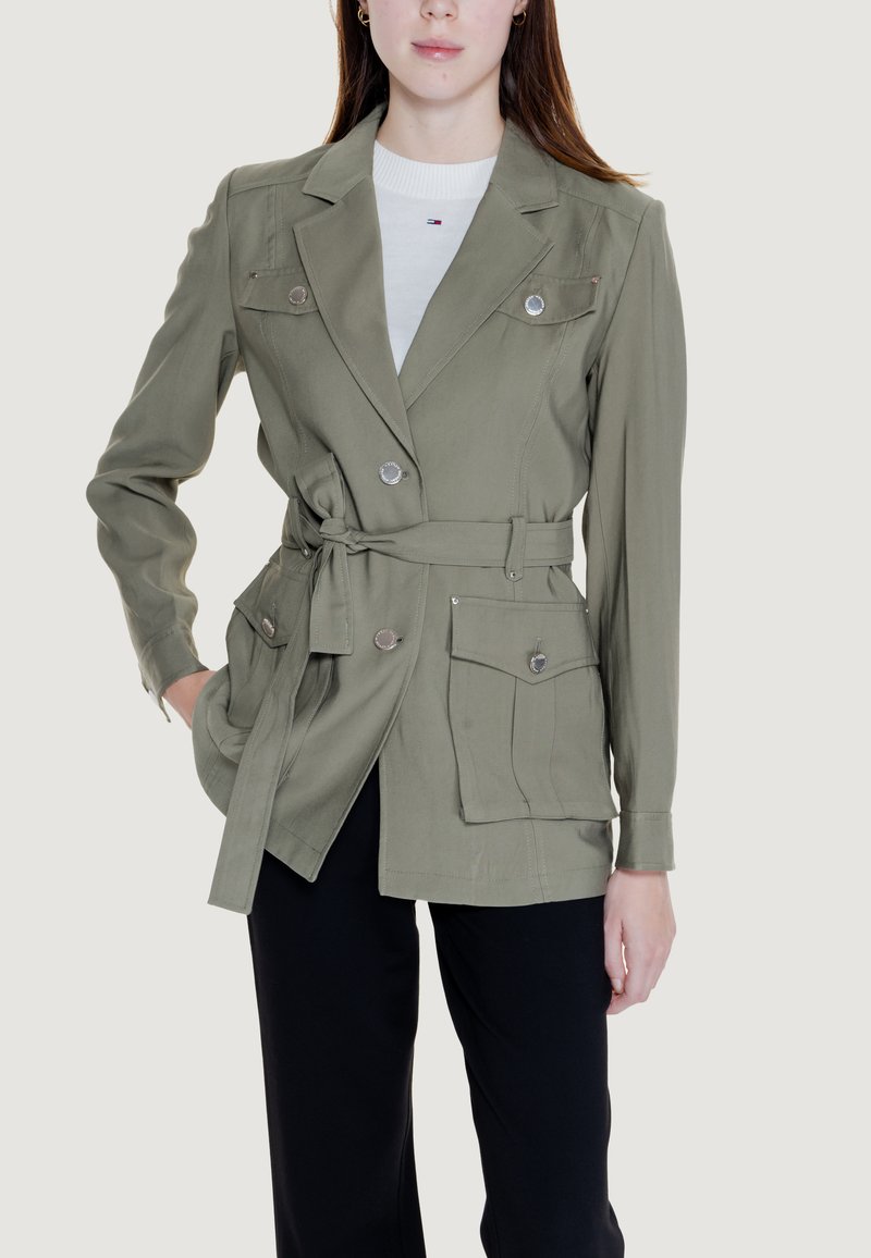 Giacca utilitaria verde oliva con vita regolabile, due tasche a toppa, dettagli in bottone argentato e maniche lunghe, abbinata a un top bianco.