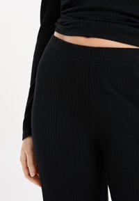 Close-up van een persoon in geribbelde zwarte broek en een bijpassend geribbeld zwart langemouwen shirt met een cropped zoom die blote huid laat zien.