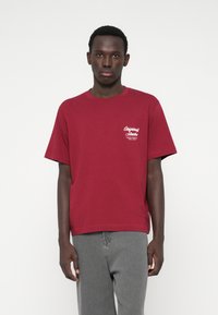 Tumši sarkans kokvilnas T-krekls ar apaļu kakla izgriezumu un īsām piedurknēm, ar baltu logo druku kreisajā krūtī.