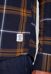 Chemise à carreaux bleu marine avec des rayures orange et bleu, tissu tissé, ourlet courbe, et une étiquette blanche portant l'inscription "TOM TAILOR".