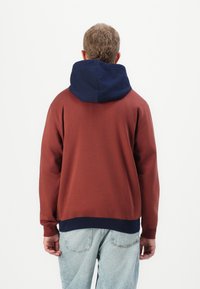Homme avec des cheveux courts et bouclés portant un sweat à capuche couleur rouille avec une capuche bleu marine et un jeans bleu clair, de dos contre un fond uni.