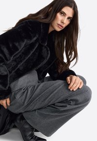 Zwarte nepbont jacket met een kraag, gecombineerd met hooggemonteerde grijze wijde pantalons en zwarte hoge hakken. Glad textuur zichtbaar.