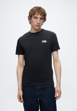 Junger Mann trägt ein schwarzes Karl Lagerfeld T-Shirt und dunkelblaue Jeans, steht vor einem einfachen weißen Hintergrund.