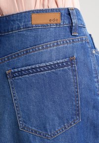 Falda de mezclilla en azul medio con una etiqueta de cuero que dice "edc". Cuenta con un bolsillo trasero con costura contrastante y una textura suave.