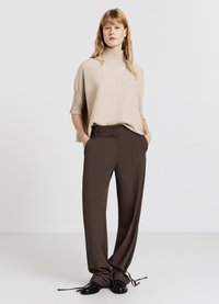 Beige kurzärmliger Strickpullover mit hohem geripptem Kragen, kombiniert mit braunen maßgeschneiderten Hosen in lockerer Passform und Kordelzugbündchen. Schwarze Schuhe.