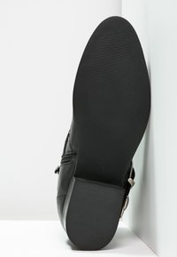 Suola di un boot in pelle nera con tacco a blocco appoggiato contro pareti bianche, che mostra una presa texture e un dettaglio della cerniera laterale.