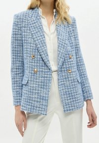 Světlomodrý tweedový blejzr s dvojitým zapínáním na knoflíky, zlatými knoflíkovými detaily a dvěma bočními kapsami, ve spojení s bílou blůzou a kalhotami.