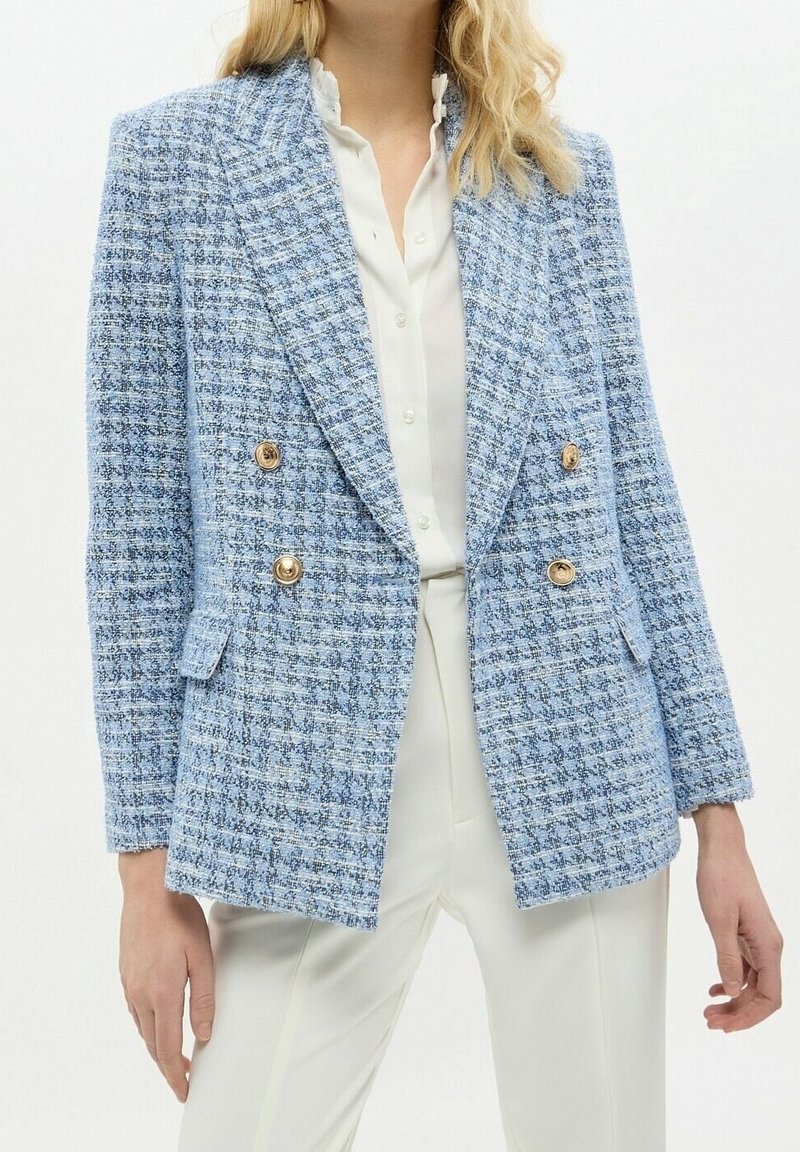 Světlomodrý tweedový blejzr s dvojitým zapínáním na knoflíky, zlatými knoflíkovými detaily a dvěma bočními kapsami, ve spojení s bílou blůzou a kalhotami.