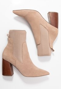 Beżowe zamszowe botki za kostkę z ostry narożnikiem, teksturowanymi elastycznymi panelami i słupkowymi drewnianymi obcasami. Posiadają pętelkę do łatwego zakładania.