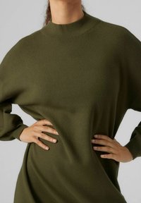 Žena oblečená v olivově zelených pletených šatech s dlouhým rukávem a vysokým výstřihem, pózuje s rukama na bocích na světlém pozadí.