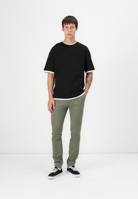 T-shirt noir à manches courtes avec un bord blanc, associé à un pantalon slim fit vert olive et des baskets noires avec des semelles blanches. Modèle debout.