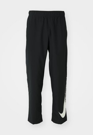 Sorte træningsbukser med elastisk talje, der taper ind, og har et stort hvidt Nike-logo på venstre ben. Blødt, letvægtsstof.