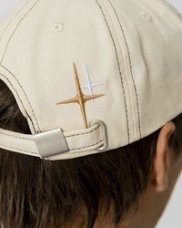 Bežs kokvilnas cepure ar regulējamu jostu, kā arī ar izšūtām zelta un baltām zvaigžņu ornamentiem aizmugurē un kontrastējošiem melniem šuvēm.