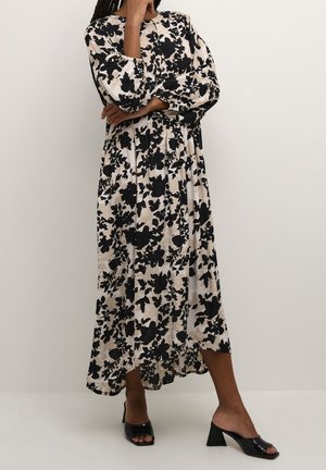 Femme debout, bras croisés, portant une longue robe fleurie noire et blanche et des mules ouvertes à talons noires sur un fond uni.