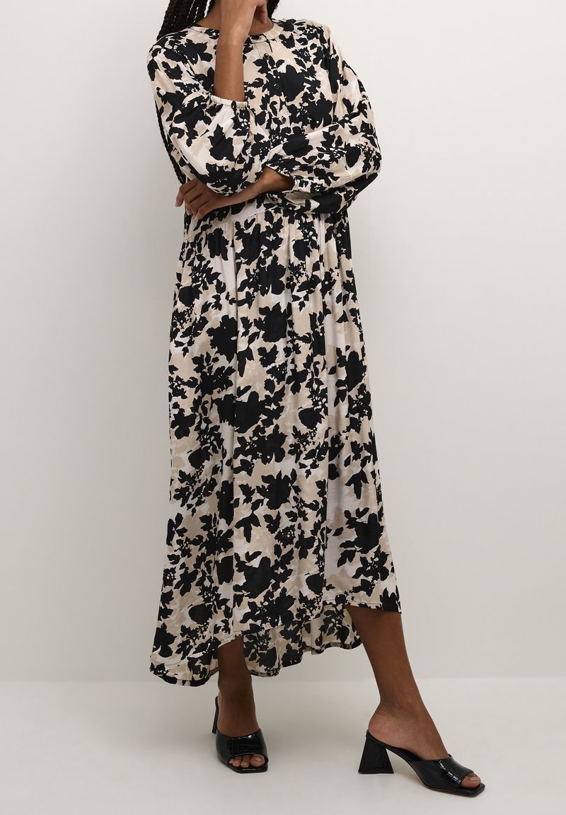 Femme debout, bras croisés, portant une longue robe fleurie noire et blanche et des mules ouvertes à talons noires sur un fond uni.
