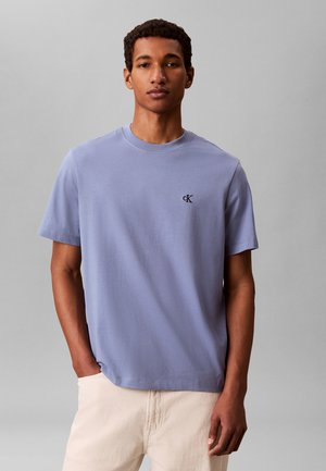 Joven con una camiseta de manga corta color violeta claro con el logo "cK" en el pecho y pantalones beige, de pie frente a un fondo gris liso.