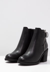 Bottes en cuir noir à cheville avec un bout rond, une fermeture éclair sur le côté et une semelle à talon épais. Dotées d'une sangle avec une boucle métallique en accent.